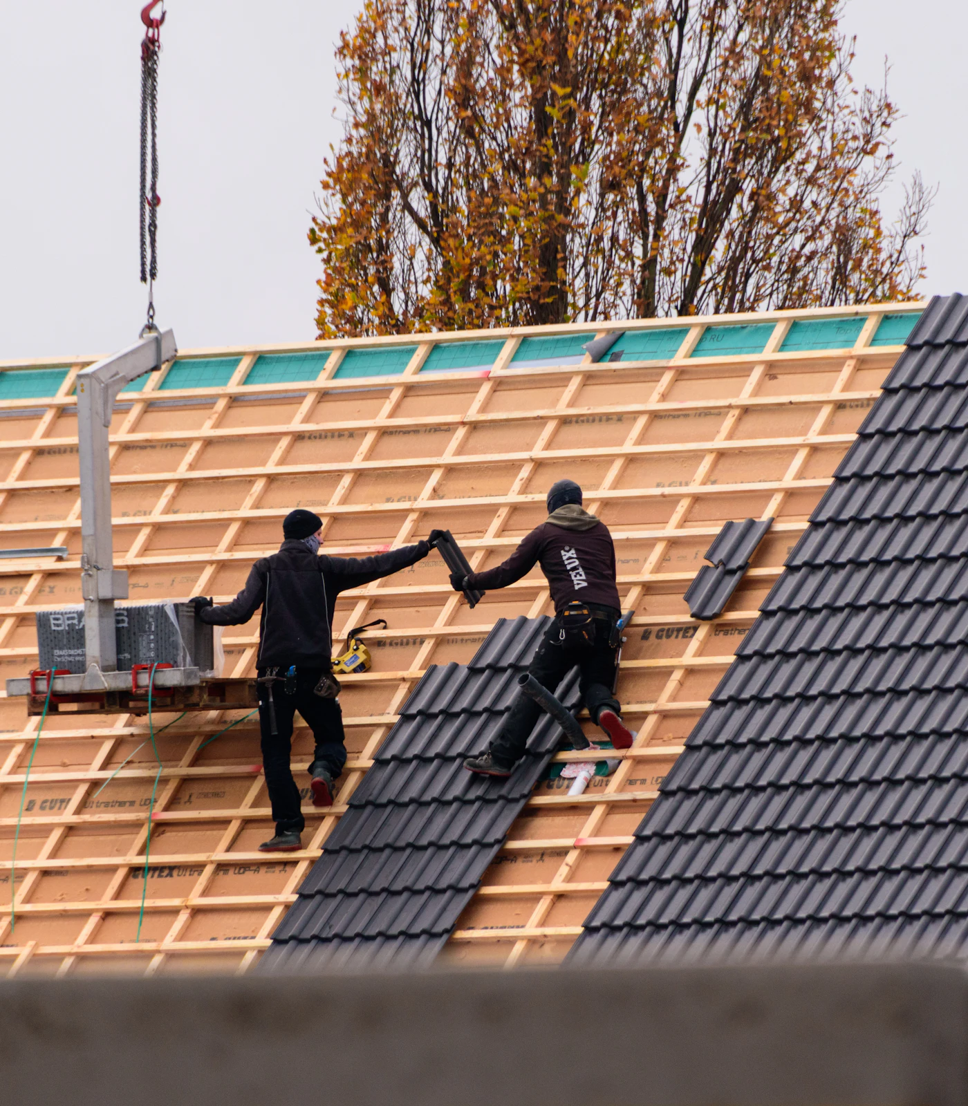 Twee dakwerkers leggen nieuwe dakpannen op een woning in opbouw, met een kraan en herfstbomen op de achtergrond